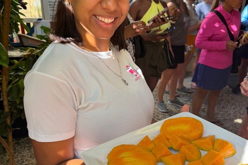 Eine Person hält einen Teller mit geschnittenem Obst, vermutlich Mangos, im Freien in Gesellschaft anderer Menschen. 5