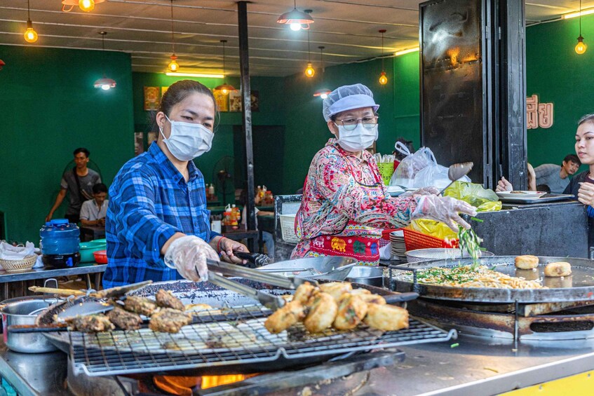 A street food vendor grilling meat and vegetables on a large grill, with customers and other vendors in the background. 5