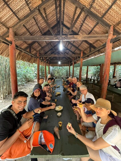 A group of people seated at a long table under a thatched roof structure, surrounded by greenery. 3