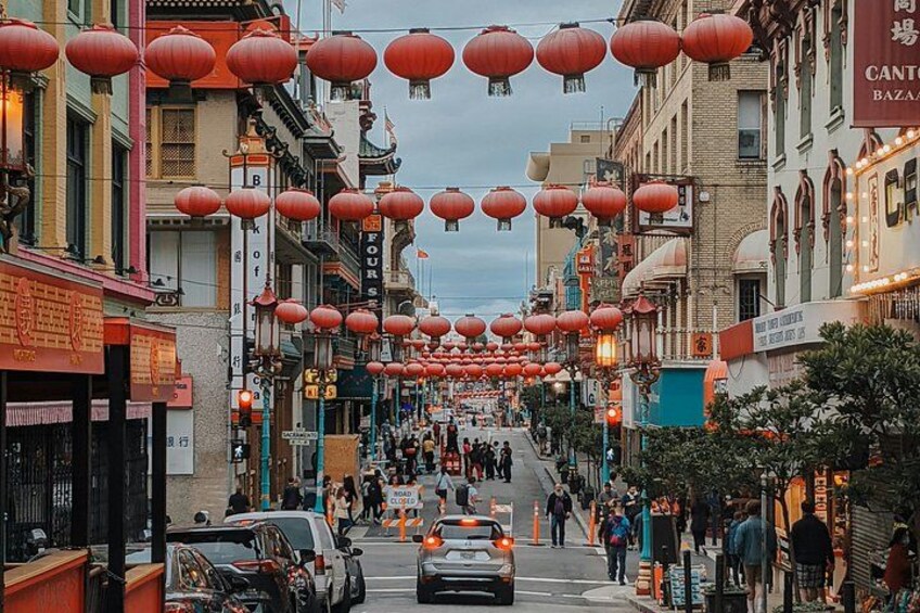 A street lined with shops and red lanterns, with people walking and cars parked along the road. 4