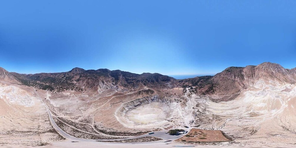 A panoramic view of a barren landscape with a winding road, a large open pit, and distant mountains under a clear blue sky. 3