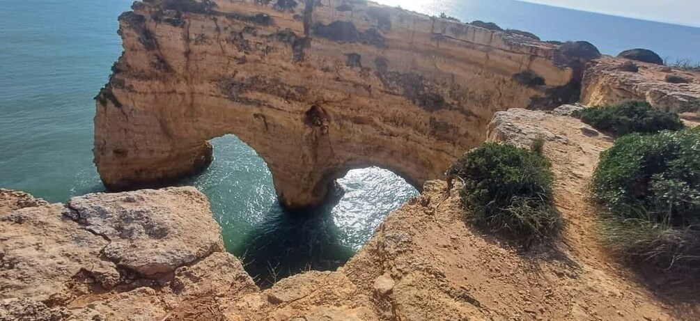 A natural rock arch formation over a body of water, surrounded by sandy terrain and sparse vegetation. 2