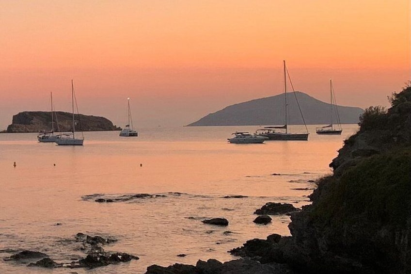 A sunset over a calm sea with several sailboats anchored, rocky coastline, and a distant hill. 5