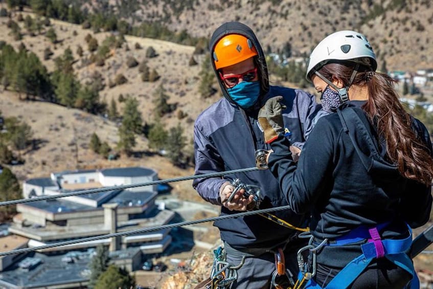 Two individuals in safety gear, one assisting the other with harness adjustments, overlooking a mountainous landscape with buildings in the background. 2