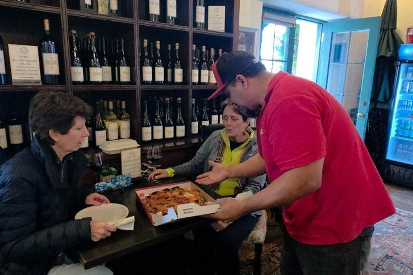 En person serverer pizza til tre personer, der sidder ved et bord i et rum med vinflasker på hylder. 3