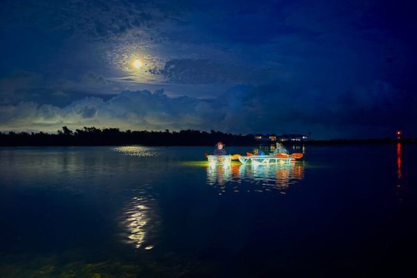 Un lago tranquilo de noche con luna llena, barcos iluminados y una orilla con edificios. 1