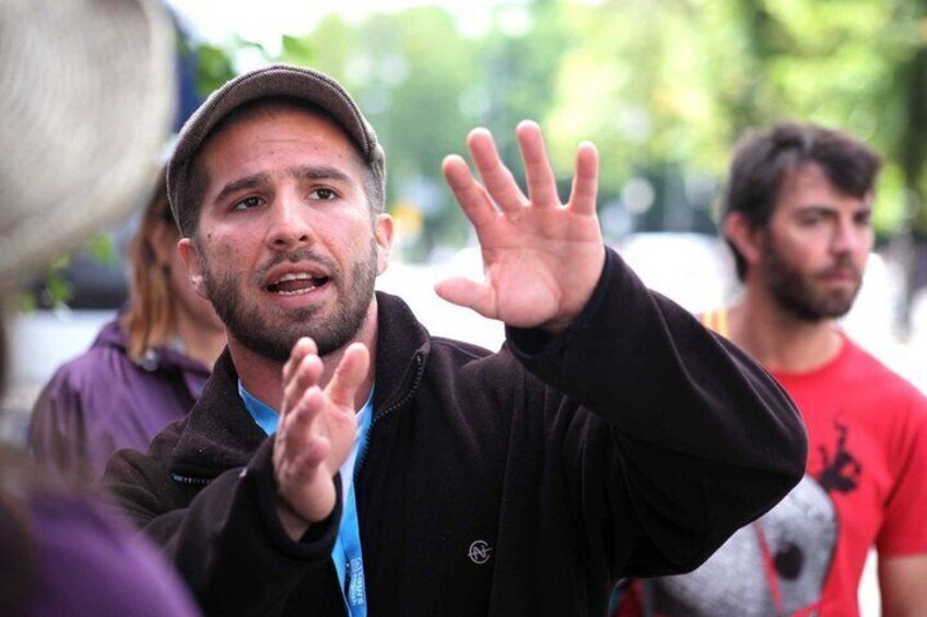 A person gesturing with their hands, wearing a dark jacket and a cap. 1
