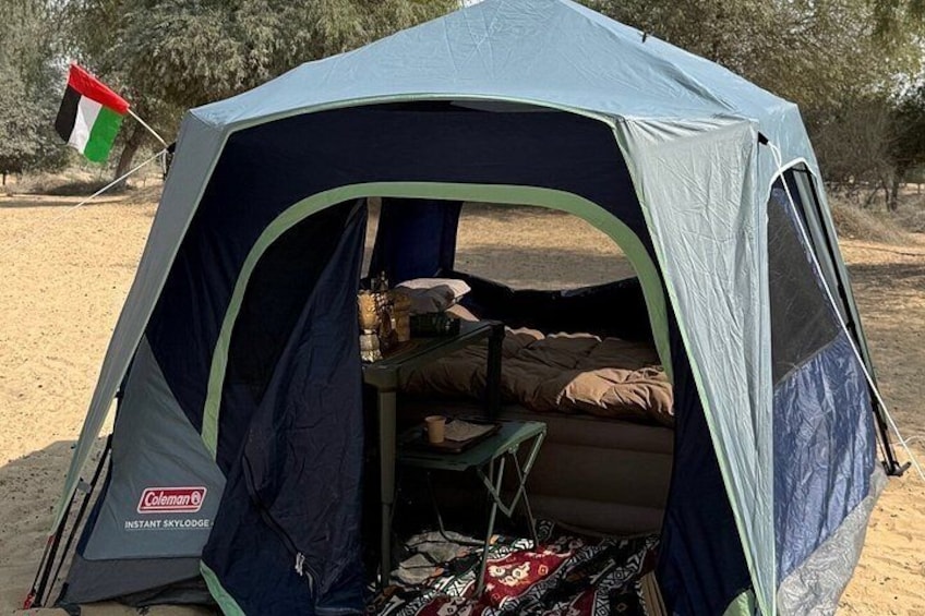Ein Coleman-Zelt ist in einer Wüstenlandschaft aufgebaut, mit einer Flagge, einem Tisch und Campingausrüstung im Inneren. 3