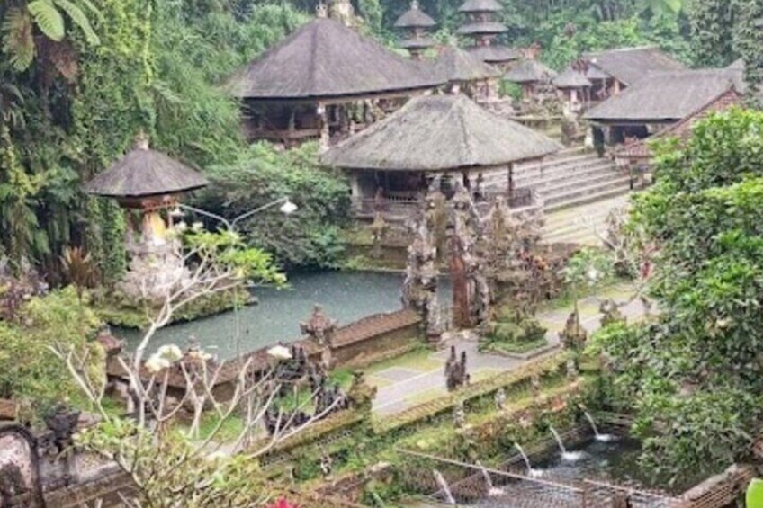 A traditional Balinese temple complex with thatched-roof structures, a central water feature, and lush greenery. 1