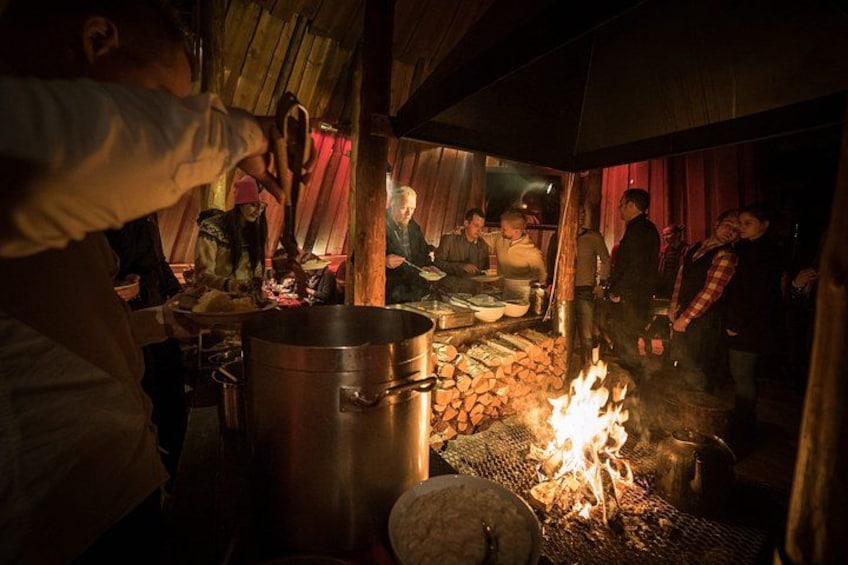 A rustic outdoor cooking scene with a large pot, a fire, and people gathered around. 4
