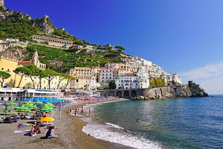 A coastal town with colorful buildings, a beach with umbrellas, and a clear blue sea. 1