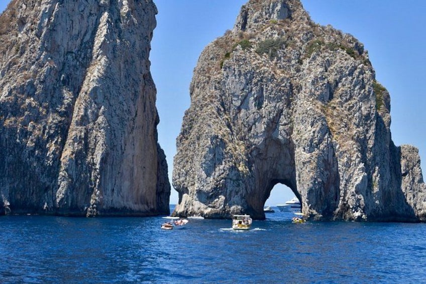 A large rock formation with a natural arch over the water, surrounded by smaller rocks and clear blue sea. 3