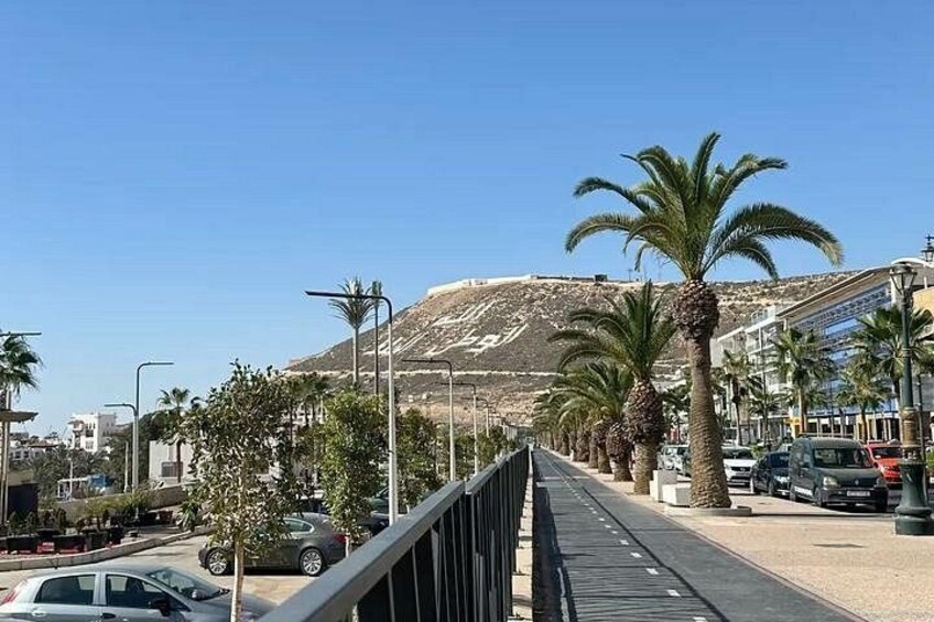 Una strada costiera con palme, una montagna sullo sfondo e un ponte pedonale. 1