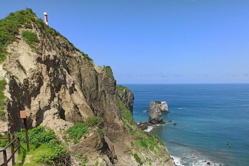 A coastal cliff with a lighthouse, rocky outcrops, and a clear blue sea. 1