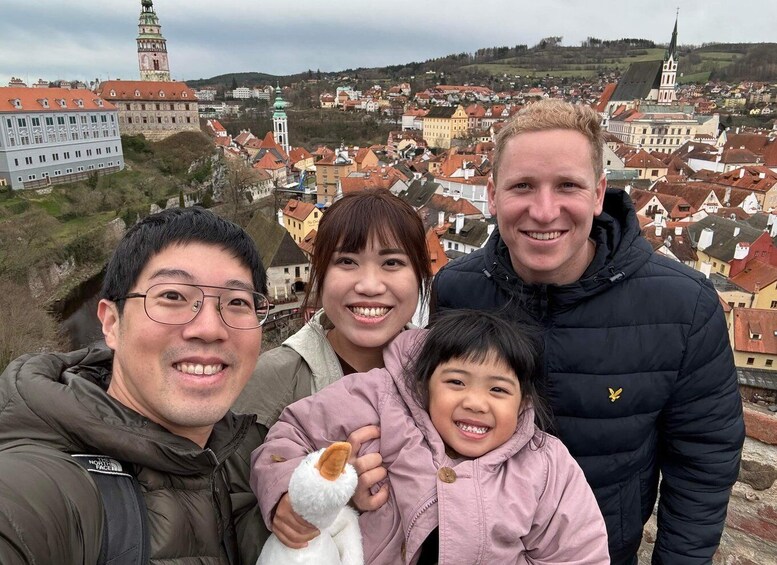 A family with a child holding a stuffed toy, standing in front of a historic town with red-roofed buildings and a prominent church tower. 4