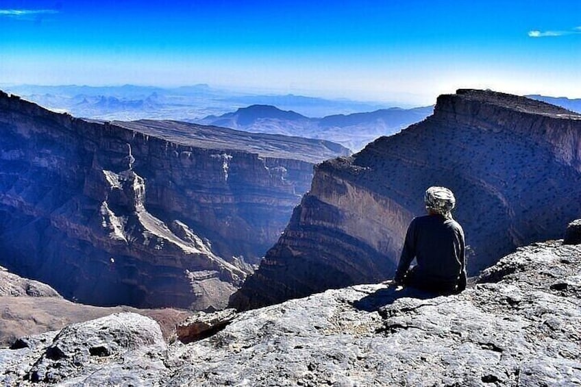 A person sitting on a rocky outcrop overlooking a deep canyon with layered rock formations. 2