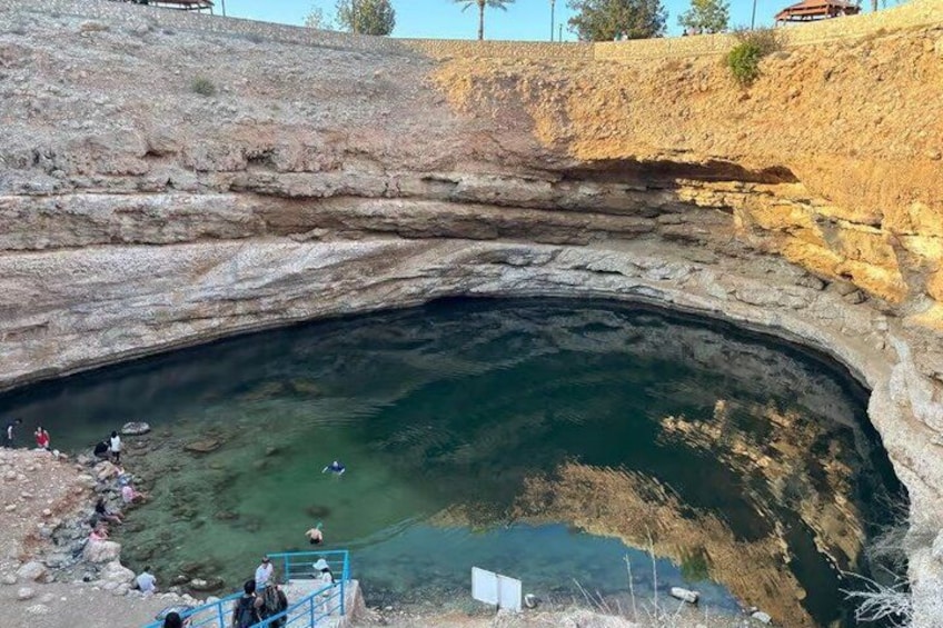 A large natural cave opening with clear water, rocky walls, and people observing from the edge. 2