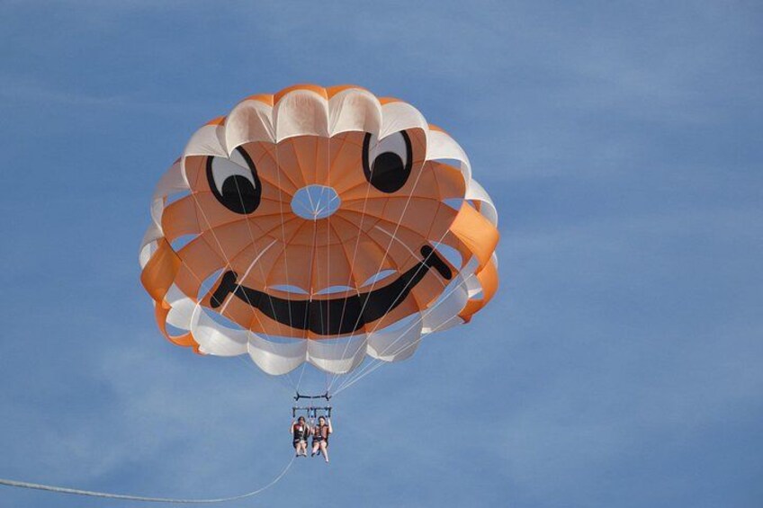 A parachute with a smiling face design, a clear blue sky, and a person harnessed to the parachute. 1