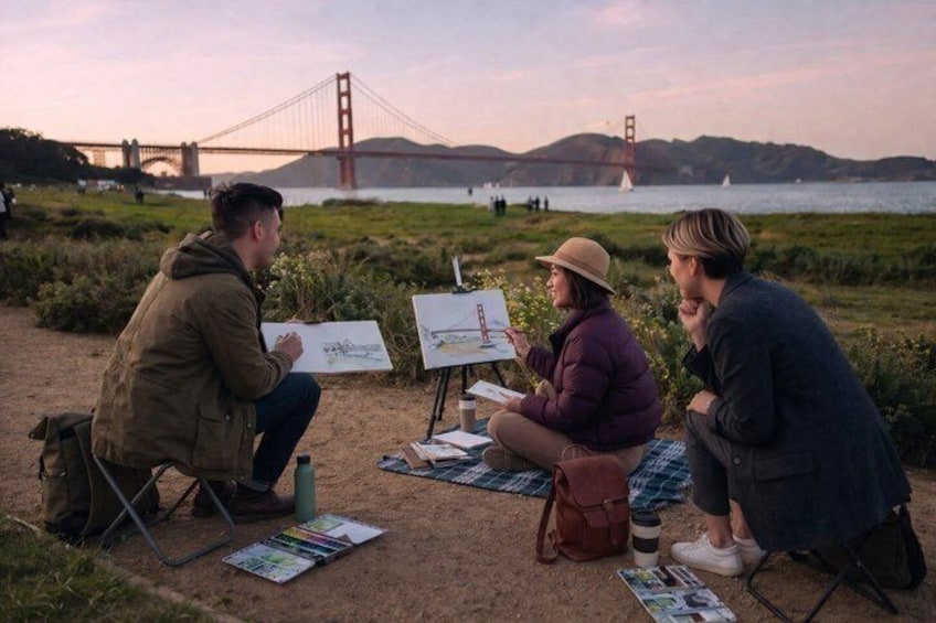 Three people sitting on a blanket by a body of water with a bridge in the background. 2