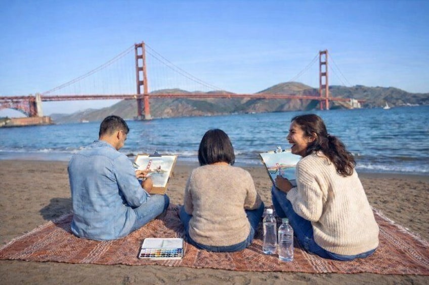 Three people sitting on a blanket by the beach with a bridge in the background. 3
