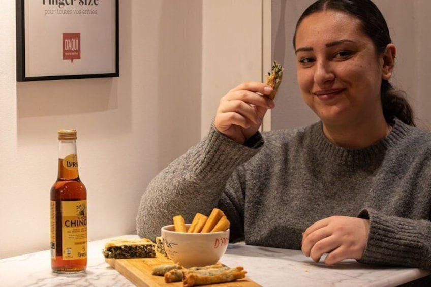 En person sidder ved et bord med en flaske Ching vin, en skål med snacks og et træbræt med mad. 1