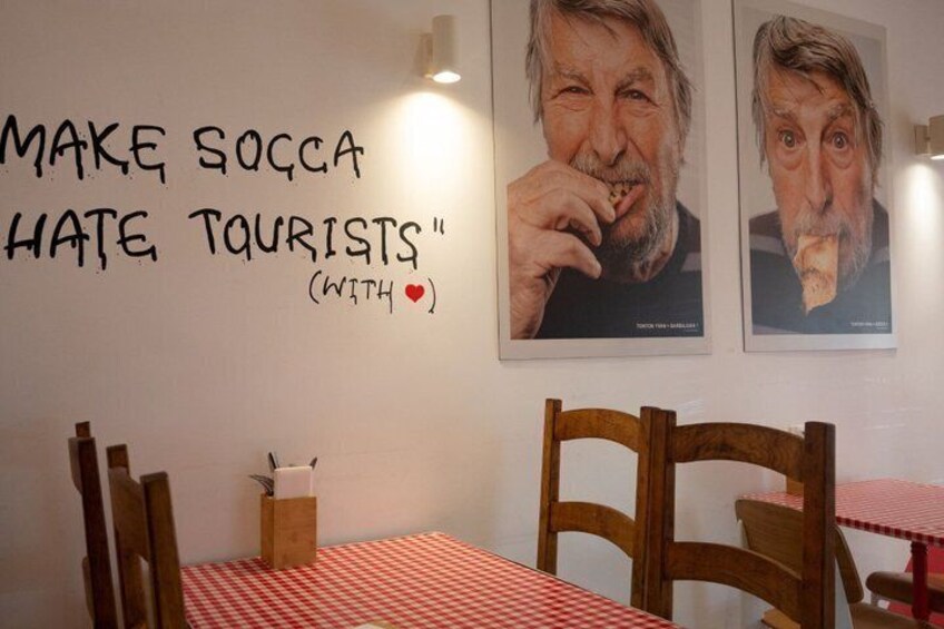 Et caféinteriør med et rødt og hvidt ternet dug, træstole og en væg med et plakat og et skilt der siger 'MAKE SOCCA HATE TOURISTS' med et rødt hjerte-symbol. 3