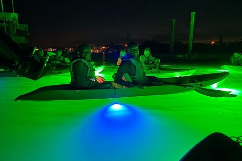 Personas sentadas en esteras con botes, bajo una iluminación verde, frente a la orilla del agua de noche. 3