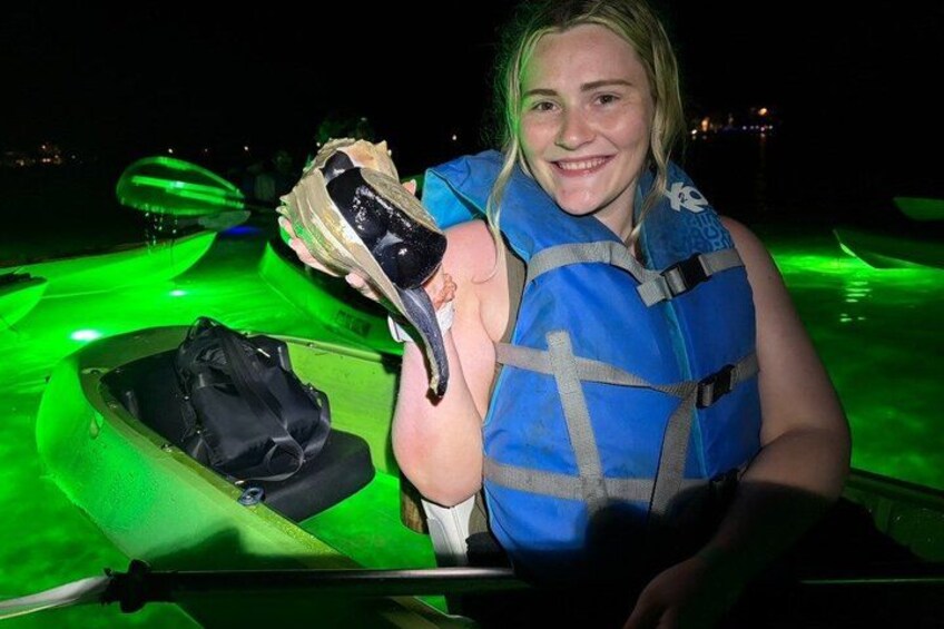 Une personne tenant une grande coquille, portant un gilet de sauvetage et se tenant à côté d’un bateau éclairé de lumière verte. 5