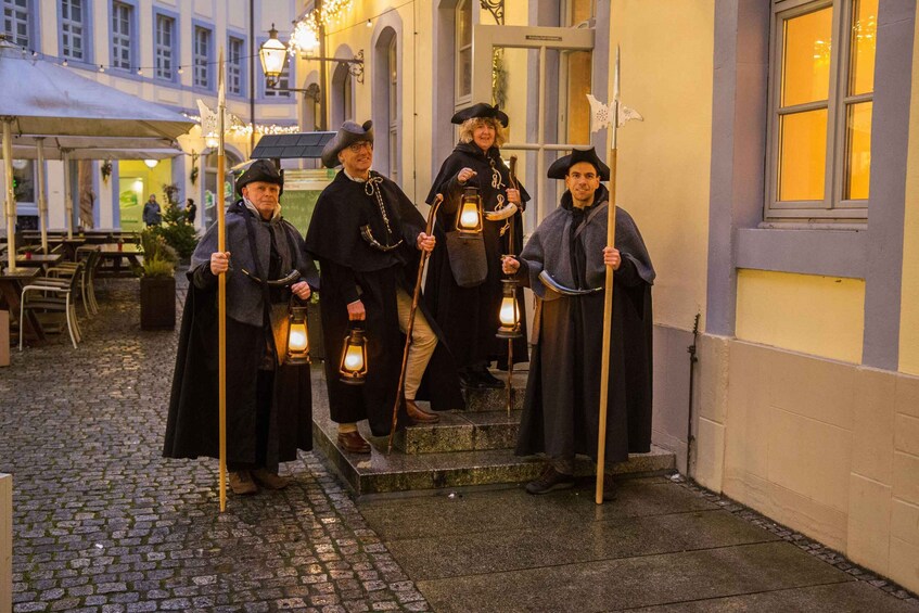 Four people in historical costumes holding lanterns, standing on steps outside a building. 1