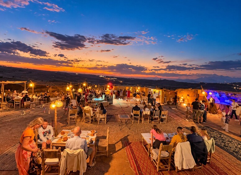 Un área de comedor al aire libre con gente sentada en mesas bajo luces colgantes, con el atardecer de fondo. 1