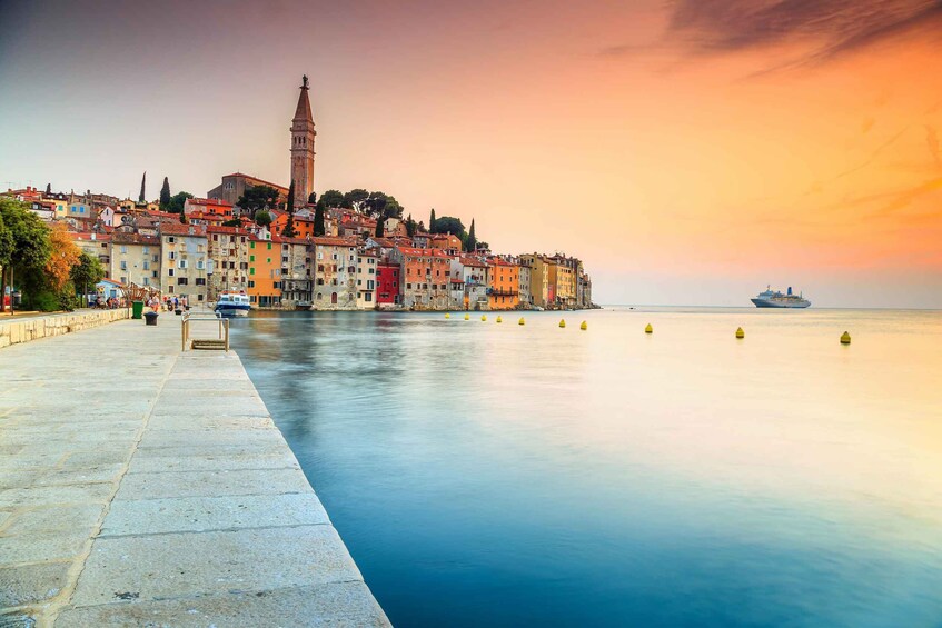 A coastal cityscape with a prominent tower, colorful buildings, and a calm sea at sunset. 1