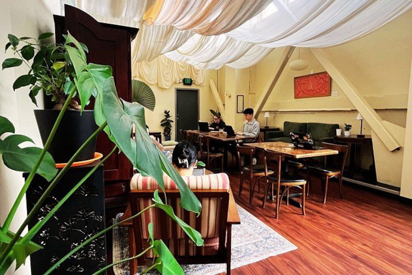A cozy restaurant with wooden tables and chairs, a large plant, and a fan on the wall. 2