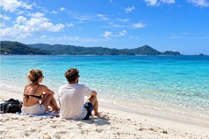 Two people sitting on a sandy beach with clear turquoise water and distant mountains. 4