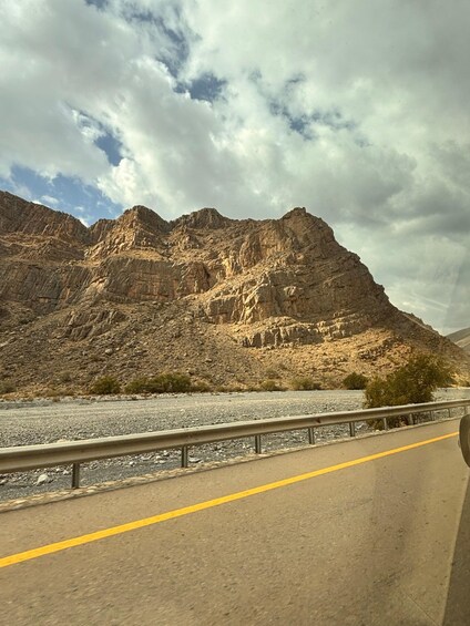 A highway with a guardrail, a river, and a rocky mountain range under a cloudy sky. 4