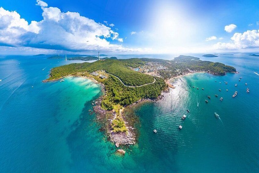 An aerial view of a coastal island with clear turquoise waters, a dense forested area, and several boats anchored near the shore. 3