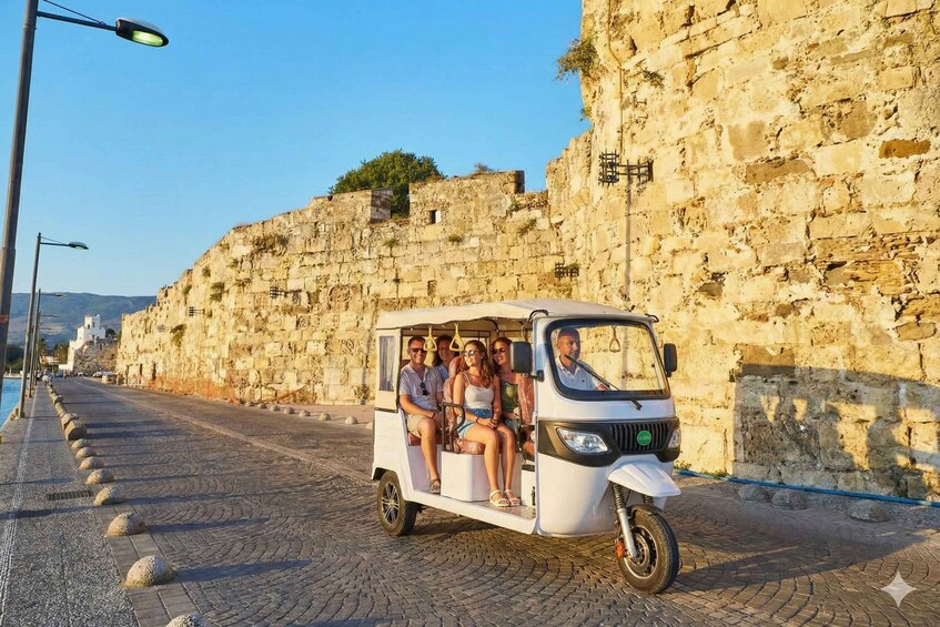 A white three-wheeled vehicle with passengers on a cobblestone street beside a stone wall. 1