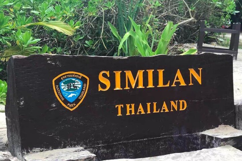 Un letrero de madera con la leyenda 'SIMILAN THAILAND' en letras doradas, un logotipo con un emblema azul y amarillo, y vegetación de fondo. 2