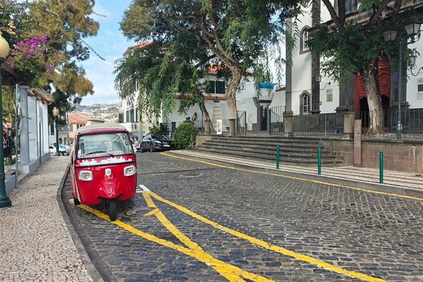 A red vintage car parked on a cobblestone street with yellow lines, surrounded by trees and buildings. 2