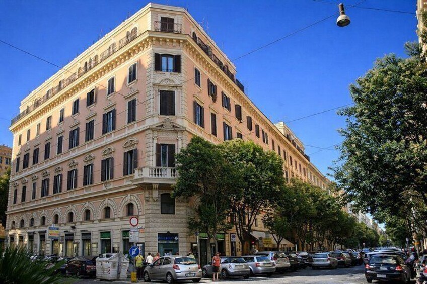 Ein mehrstöckiges Gebäude mit klassischer Architektur, eine Straße mit geparkten Autos und Bäumen entlang des Bürgersteigs. 5