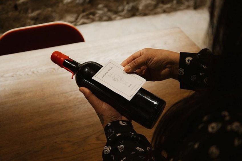 A person holding a wine bottle with a label, seated at a wooden table. 2