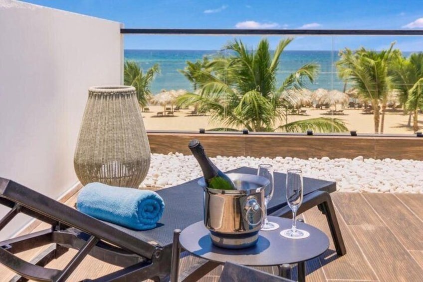 Un balcón con una mesa, una botella de champán y una copa, con vistas a una playa con palmeras y el océano. 3