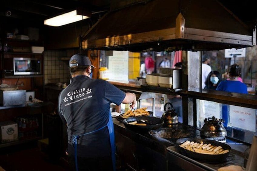 Une personne prépare des plats dans un stand de restauration de rue. On peut voir une pancarte derrière elle et une bouilloire sur le comptoir. 3