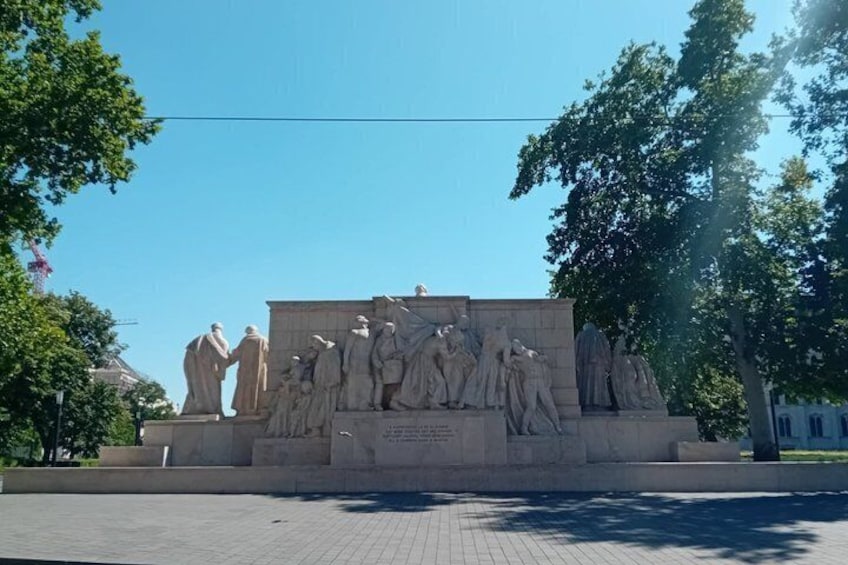 A large outdoor sculpture with multiple figures, surrounded by trees and a paved area. 2