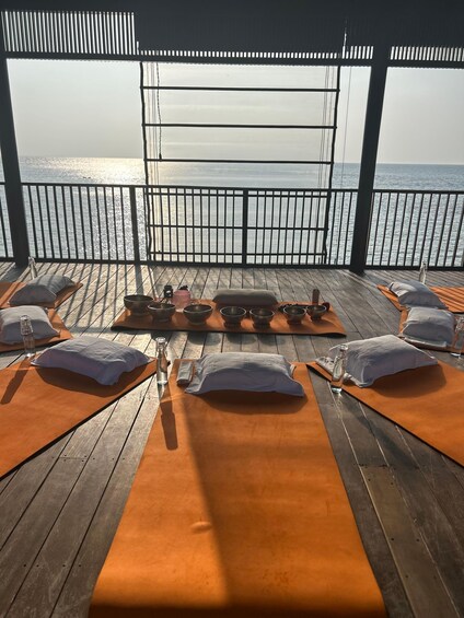 A wooden deck with yoga mats and pillows, a meditation area with bowls and a water bottle, and a view of the sea. 1