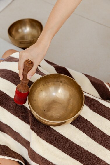 A person holding a mallet next to a large brass bowl, likely used for meditation or gong playing, on a striped fabric. 4