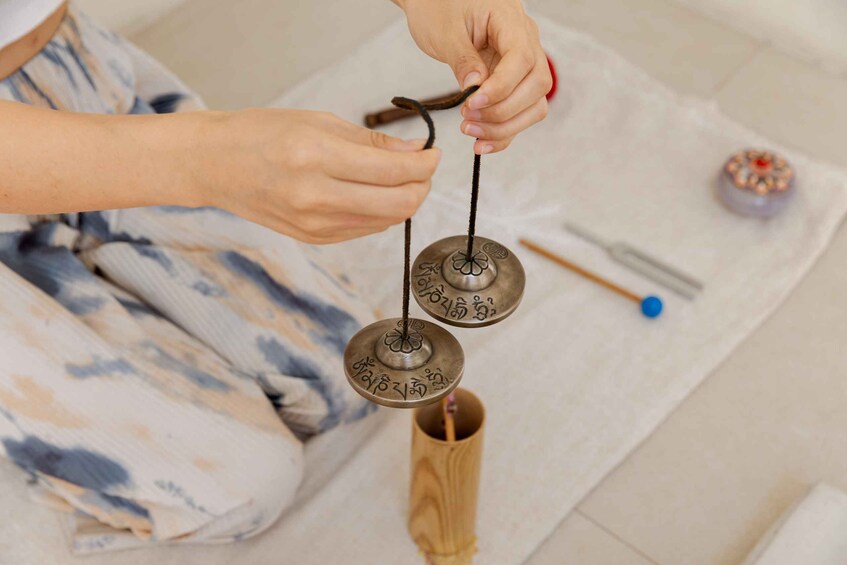 A person holding a string between two metal objects with inscriptions, sitting on a mat with a wooden container and a blue object nearby. 3
