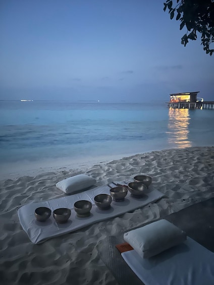 A beach with a row of bowls and pillows on a mat, a pier in the background, and calm sea. 5