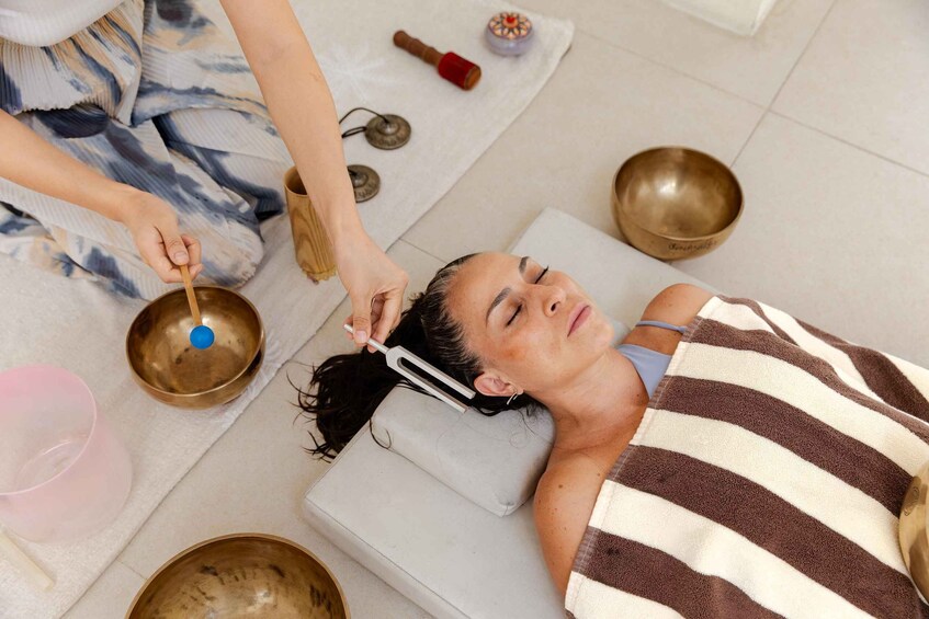 A person lying on a massage table with a striped towel, surrounded by various bowls and a person performing a massage. 1