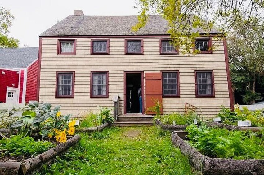 A two-story house with a red door and windows, surrounded by a garden with various plants and a wooden fence. 3