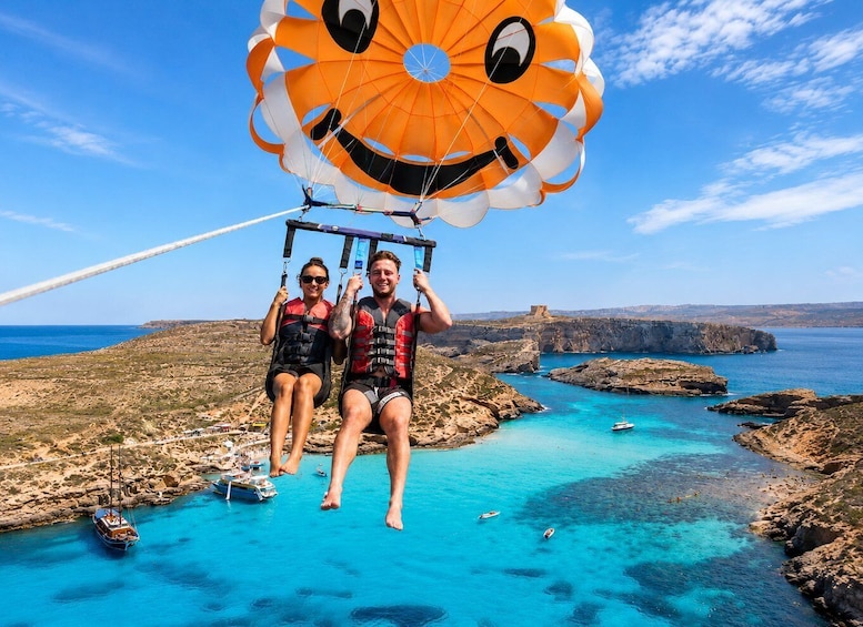 Two people on a parasail over a coastal landscape with clear blue waters and rocky terrain. 1